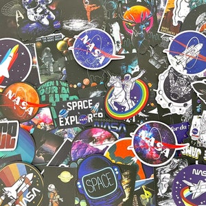 May include: A selection of space-themed stickers with diverse designs. The stickers depict rockets, planets, astronauts, and the NASA logo. Some stickers have text such as "SPACE EXPLORED" and "SPACE JAMS". The colour scheme includes blues, oranges, and purples.
