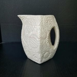 May include: White ceramic pitcher with a floral embossed design. The pitcher has a square base and a wide mouth.