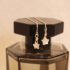 Puede incluir: Un par de pendientes de plata con colgantes en forma de estrella blanca. Los pendientes se muestran en una botella negra.