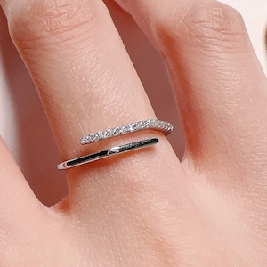 Puede incluir: Un anillo de plata con una banda fina y una fila de pequeñas piedras transparentes en la parte superior. El anillo está envuelto alrededor del dedo.