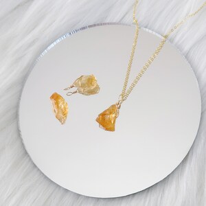 May include: A gold chain necklace with a raw, yellow citrine pendant. The necklace is displayed on a white surface with two smaller raw citrine earrings.