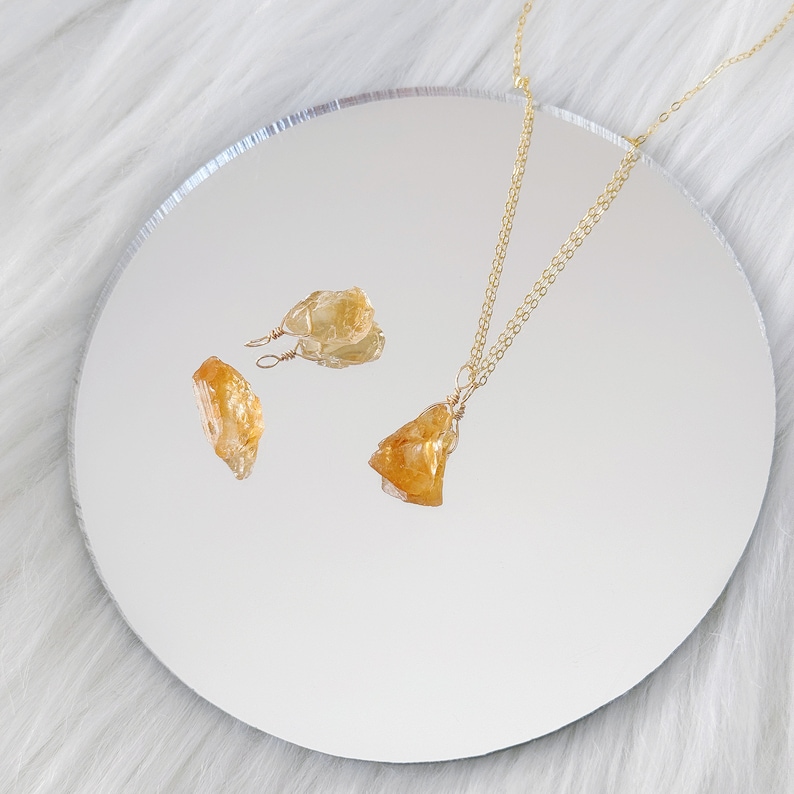 May include: A gold chain necklace with a raw, yellow citrine pendant. The necklace is displayed on a white surface with two smaller raw citrine earrings.