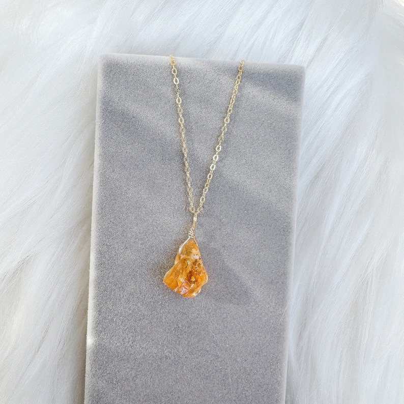 May include: A gold chain necklace with a raw, orange carnelian pendant. The pendant is wrapped in gold wire and hangs from a delicate gold chain.