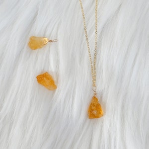 May include: A gold chain necklace with a raw, orange-brown crystal pendant. Two additional raw crystals are shown on a white fuzzy surface.
