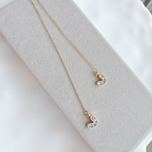 Op de afbeelding: Fijne goudkleurige ketting met twee hartvormige hangers. Elk hart is bezet met kleine, heldere kristallen. De ketting wordt gepresenteerd op een lichtgrijze ondergrond, wat een contrast creëert.