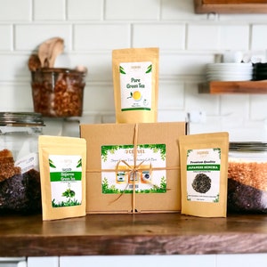 May include: A brown cardboard box with a green ribbon and a label that says "Super Green Tea Selection Gift Box". The box is surrounded by three bags of green tea, each with a different label. The labels say "Pure Green Tea", "Supreme Green Tea", and "Premium Quality Japanese Sencha".