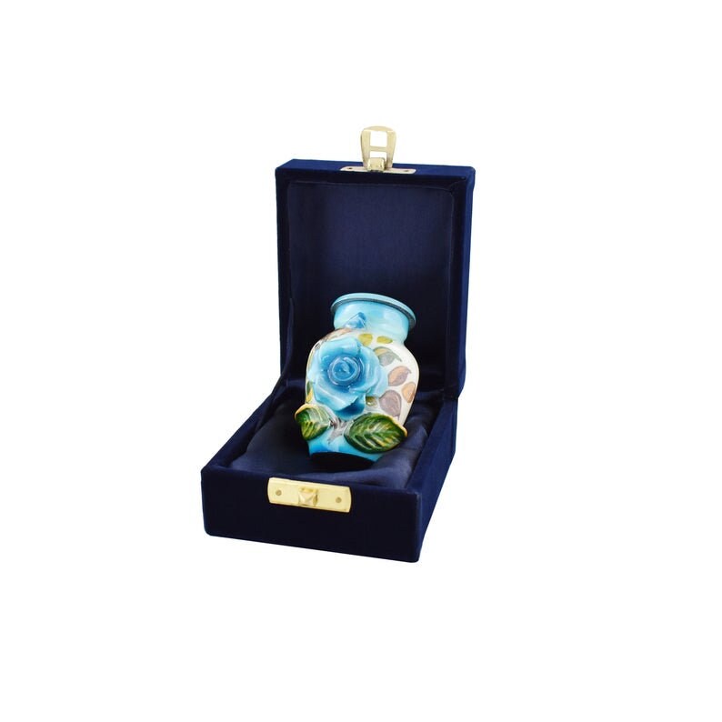 K&ouml;nnte beinhalten: Eine kleine blaue und wei&szlig;e Keramikurne mit einem floralen Design, das eine blaue Rose und gr&uuml;ne Bl&auml;tter zeigt. Die Urne befindet sich in einer blauen Samtbox mit einem goldenen Verschluss.