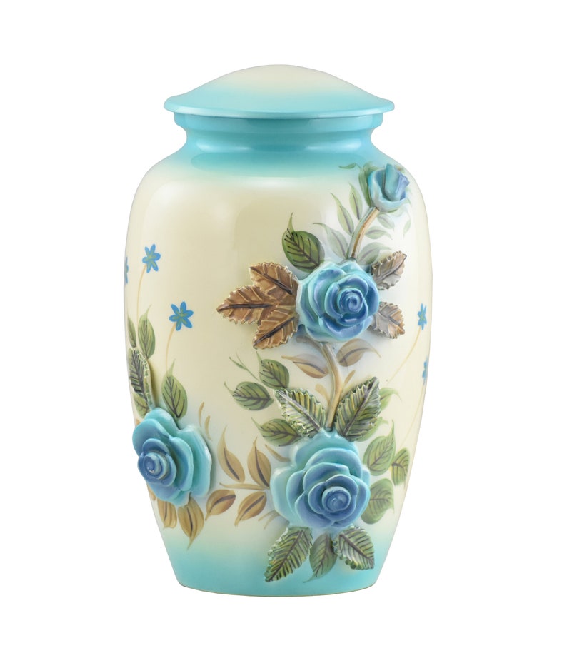 K&ouml;nnte beinhalten: Eine wei&szlig;e Keramikurne mit einem blauen Deckel und einem Blumendesign mit blauen Rosen und gr&uuml;nen Bl&auml;ttern. Die Urne hat einen Farbverlauf von hellblau zu wei&szlig;.