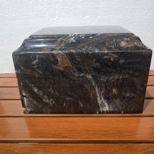 May include: A rectangular cremation urn crafted from polished stone featuring a dark brown, black, and white marbled pattern. The urn has a stepped lid design with smooth, reflective surfaces and a solid, heavy appearance with natural veining.