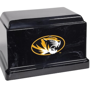 May include: A black, rectangular urn with a glossy finish. The urn features a gold and black logo of a tiger's head on the front. The urn has a lid and a base, and is designed to hold cremated remains.