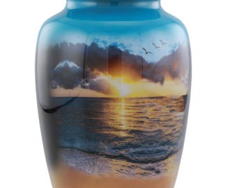 Puede incluir: Urna funeraria azul con una escena de playa al atardecer. La urna presenta una tapa azul cielo y un acabado brillante. La imagen representa una puesta de sol sobre el océano con pájaros.