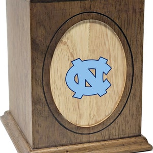 K&ouml;nnte beinhalten: Eine Holzkremierungsurne mit einer hellbraunen Oberfl&auml;che und einer hellbraunen ovalen Einlage mit einem blauen "NC"-Logo.