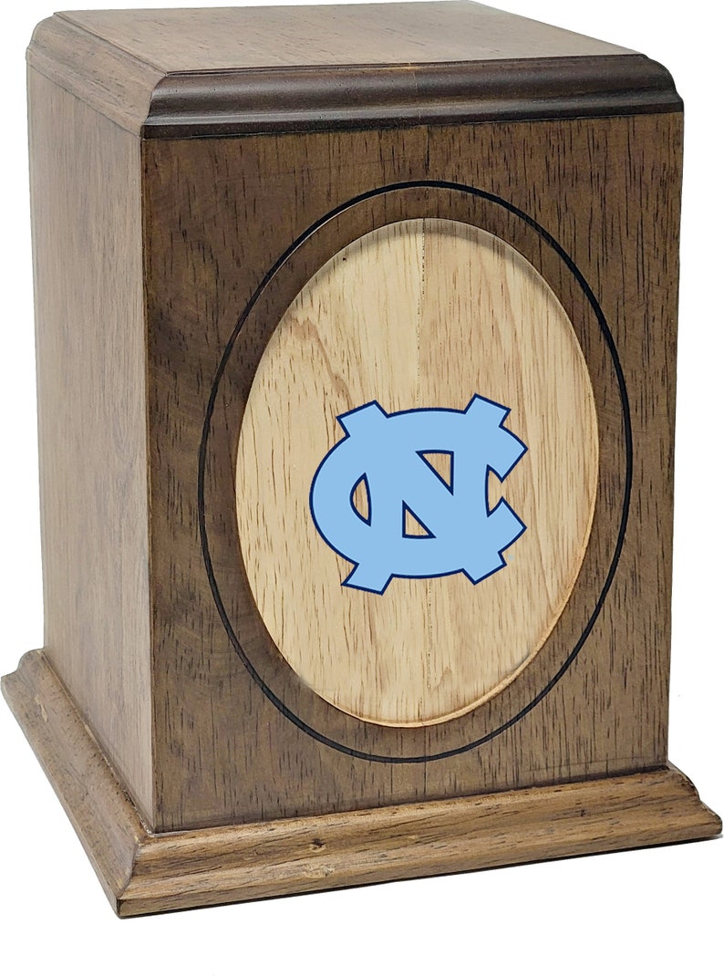 K&ouml;nnte beinhalten: Eine Holzkremierungsurne mit einer hellbraunen Oberfl&auml;che und einer hellbraunen ovalen Einlage mit einem blauen "NC"-Logo.