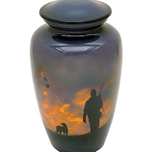 Op de afbeelding: Donkerblauwe crematieurn met deksel, met een zonsondergangscène met een jager, hond en vliegende vogels. De urn heeft een glanzende afwerking en een afgeronde vorm.