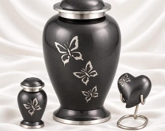 Puede incluir: Una urna funeraria de color gris oscuro con tapa y base plateadas, decorada con tres mariposas grabadas. A su lado hay una urna pequeña a juego y un recuerdo en forma de corazón sobre un soporte de metal plateado.