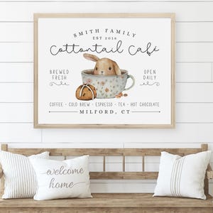 May include: A white sign with a brown wooden frame, advertising a coffee shop called "Cottontail Cafe" with a cartoon bunny in a teacup. The sign reads "Smith Family Est 2016 Cottontail Cafe Brewed Fresh Open Daily Coffee Cold Brew Espresso Tea Hot Chocolate Milford, CT"