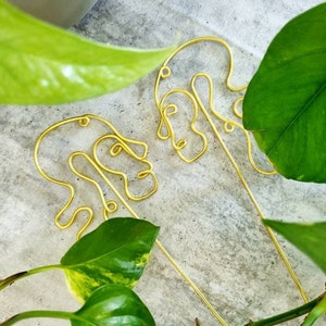 May include: Two gold wire sculptures of abstract faces, one on top of the other, against a background of green leaves.