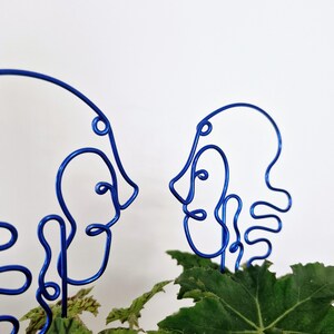 May include: Two blue wire sculptures of faces, facing each other, are placed in a pot of green leafy plants.