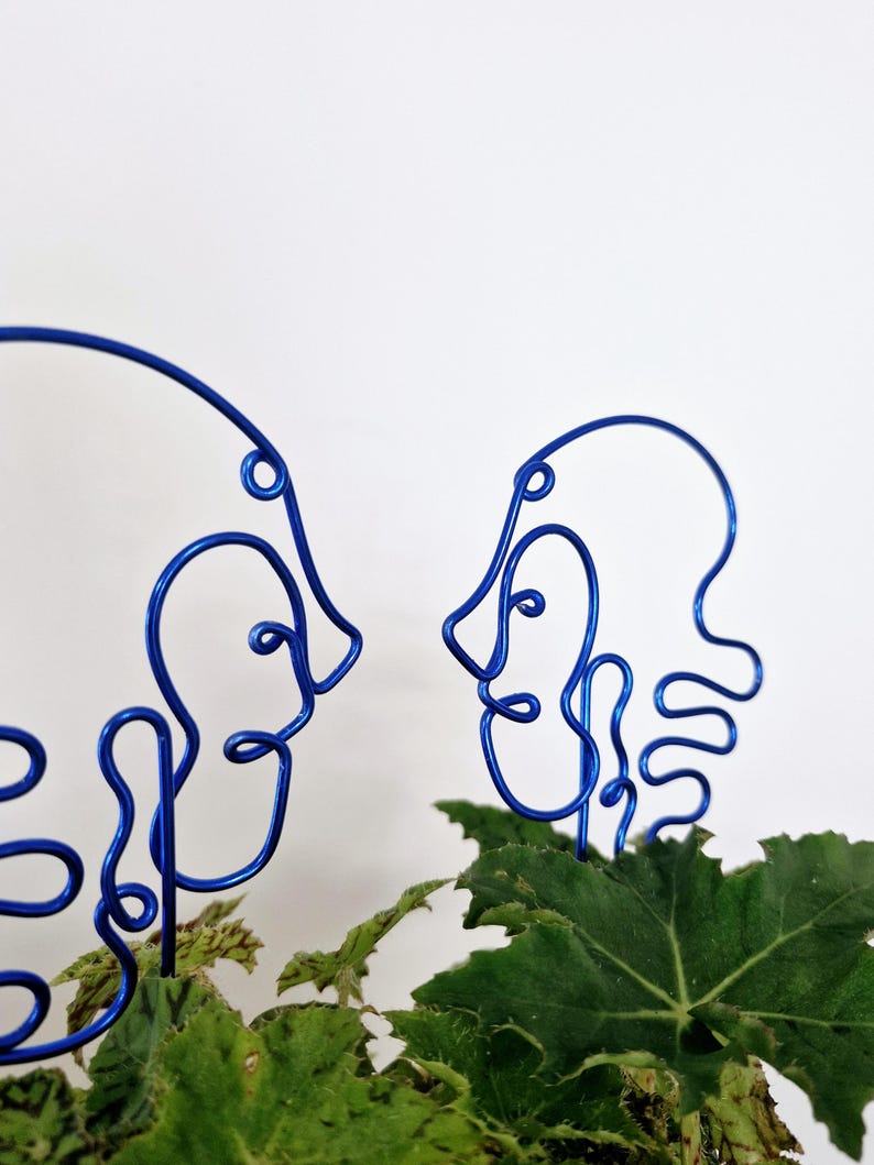 May include: Two blue wire sculptures of faces, facing each other, are placed in a pot of green leafy plants.