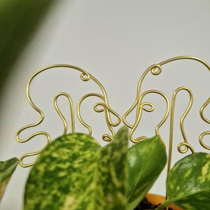 May include: Gold wire plant stakes shaped like abstract faces, used to support a potted plant with green and yellow leaves.