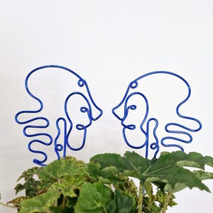 May include: Two blue wire sculptures of abstract faces facing each other. The sculptures are placed in a pot of green leafy plants.