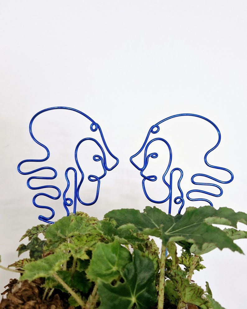 May include: Two blue wire sculptures of abstract faces facing each other. The sculptures are placed in a pot of green leafy plants.