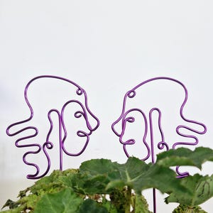 May include: Two purple wire sculptures of abstract faces facing each other. The sculptures are placed in a green leafy plant.