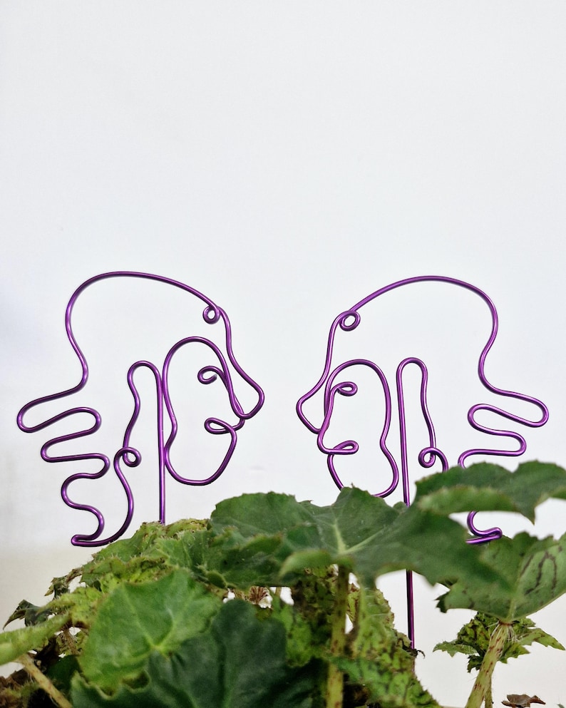 May include: Two purple wire sculptures of abstract faces facing each other. The sculptures are placed in a green leafy plant.