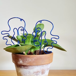 May include: Two blue wire sculptures of faces are placed in a terracotta pot with a green plant. The pot has a white glaze and is sitting on a wooden surface.