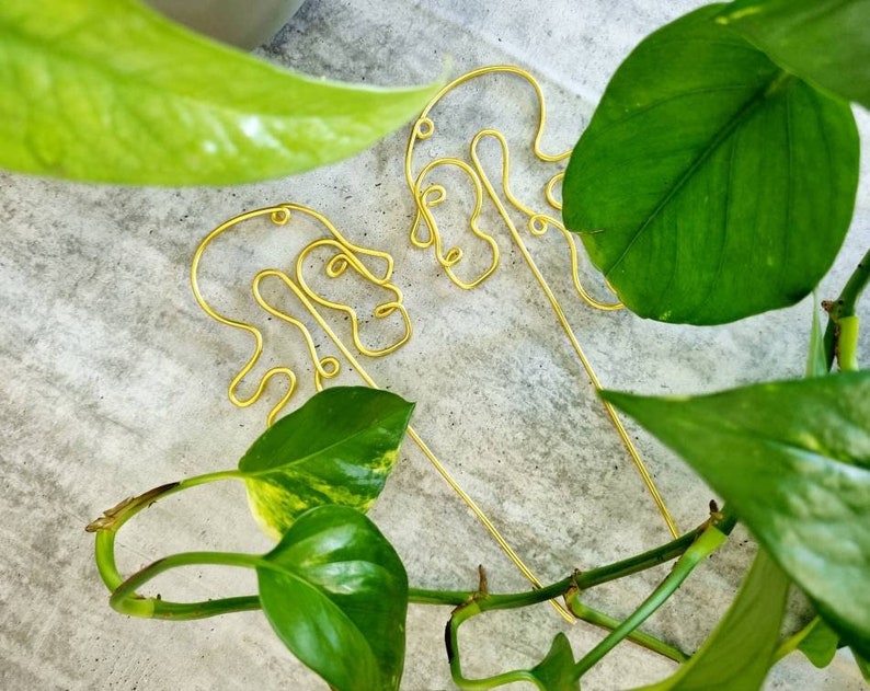 May include: Two gold wire sculptures of abstract faces, one on top of the other, against a background of green leaves.