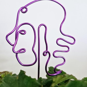 May include: Purple wire sculpture of a face with abstract hair. The sculpture is placed in a pot of green plants.