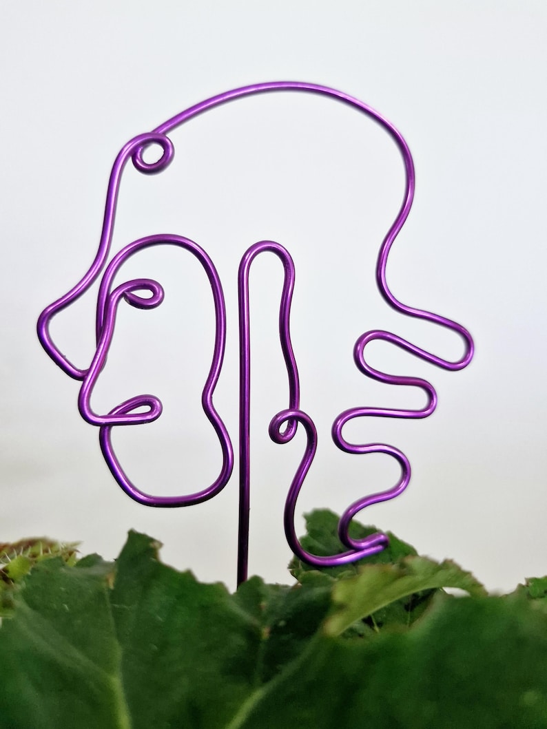 May include: Purple wire sculpture of a face with abstract hair. The sculpture is placed in a pot of green plants.