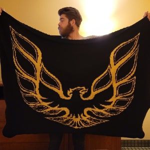 Könnte beinhalten: Eine schwarze gestrickte Wolldecke mit einem goldenen gestickten Phönix-Design. Die Decke wird von einer Person gehalten.