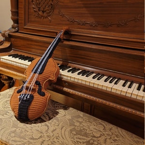 Könnte beinhalten: Eine gehäkelte Violine in Braun und Schwarz, die auf einem hölzernen Klavierhocker mit einem floralen Stoffbezug liegt.