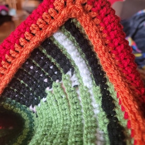 May include: Close-up of a crocheted blanket with a red, orange, and green striped border. The blanket is made with a variety of colors, including black, white, and green.
