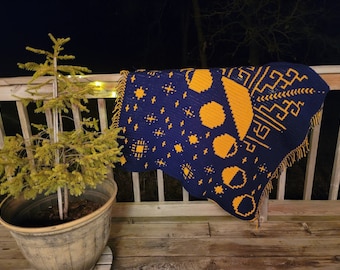 Puede incluir: Una manta de crochet azul y amarilla con un patrón geométrico de lunas y estrellas. La manta está drapeada sobre una barandilla de madera.