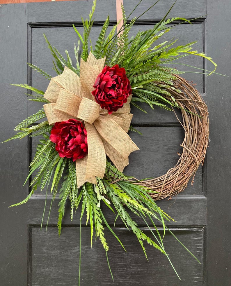 May include: A grapevine wreath decorated with a burlap bow and two red peonies. The wreath is surrounded by green foliage.