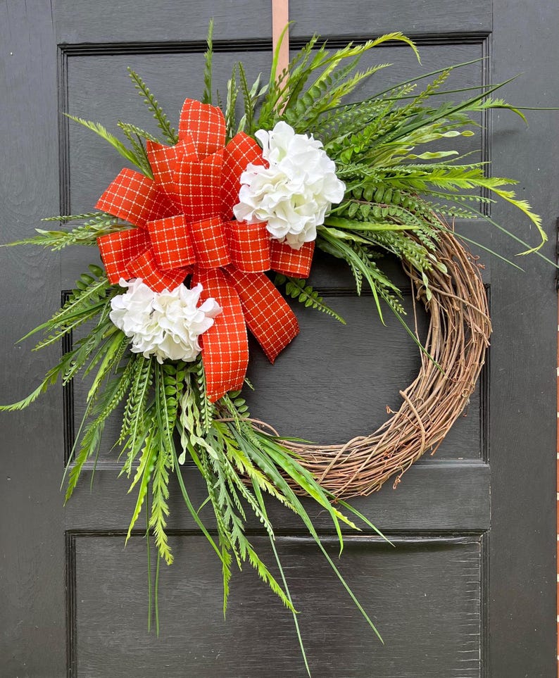May include: A grapevine wreath decorated with green foliage, white hydrangea flowers, and a large red and white plaid bow.