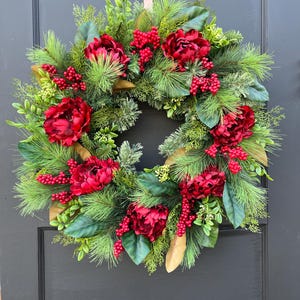 May include: A festive wreath featuring vibrant red peonies, berries, and lush green foliage. The wreath is adorned with pine needles and magnolia leaves, creating a full and textured appearance. It is designed for door decor.