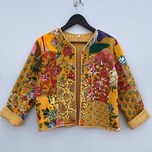 May include: A yellow quilted jacket with a floral patchwork design. The jacket has a V-neck and long sleeves with striped cuffs.