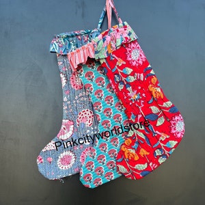 May include: Two quilted Christmas stockings with floral patterns in shades of red, pink, blue, and green. The stockings are hanging from a hook on a dark gray wall.