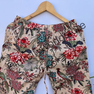 May include: Beige floral print pajama pants with a drawstring waist and white trim. The pants feature a vibrant pattern of red, teal, and green flowers, birds, and foliage. The pants are hanging on a wooden hanger.