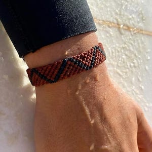 Könnte beinhalten: Ein gewebtes Armband mit einem sich wiederholenden geometrischen Zickzackmuster aus dunkelbraunem und schwarzem Garn. Das Armband besteht aus fest geknüpfter Kordel und liegt sicher am Handgelenk an, was ein strukturiertes und langlebiges Design zeigt.