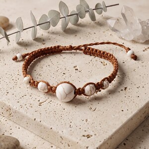 Puede incluir: Una pulsera de cordón marrón con una cuenta central grande de color blanco con patrón de mármol, flanqueada por cuatro cuentas más pequeñas a juego, con dos cuentas blancas adicionales en los extremos ajustables del cordón, hecha con nudos.