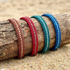 Könnte beinhalten: Vier geflochtene Armbänder in Braun-, Rot-, Türkis- und Blautönen liegen auf einem Stück Treibholz. Die Armbänder haben ein gewebtes Design und werden an einem Sandstrand präsentiert, was ein Küsten- oder Boho-Thema suggeriert.
