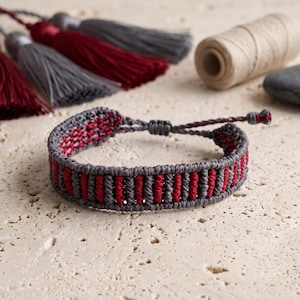 Puede incluir: Una pulsera de macramé con un patrón repetitivo de cordón gris oscuro y rojo intenso. La pulsera tiene un cierre de nudo deslizante ajustable y está construida con hilo tejido, mostrando un diseño de nudos cilíndricos texturizados.
