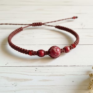 Op de afbeelding: Bordeaux macramé armband met rode kralen. De armband heeft een grote rode centrale kraal met een wit patroon, geflankeerd door kleinere rode kralen. De verstelbare sluiting zorgt voor een comfortabele pasvorm.