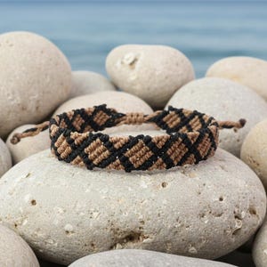 May include: A woven macrame bracelet featuring a repeating diamond pattern in alternating black and tan thread. The bracelet is constructed with a series of tight knots and includes two thin braided ties extending from each side for closure.