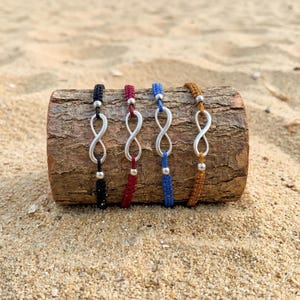 May include: Four cord bracelets featuring silver-toned infinity symbols. Each bracelet has a different coloured cord: black, red, blue, and mustard yellow. Small silver-toned beads are positioned above and below each infinity charm on the cords.