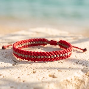 Peut inclure: Un bracelet en cordon tressé composé d'une rangée centrale de nœuds beige clair encadrée par deux rangées de nœuds rouge vif. Le bracelet présente des cordons de serrage réglables de chaque côté, terminés par de petits nœuds aux extrémités.